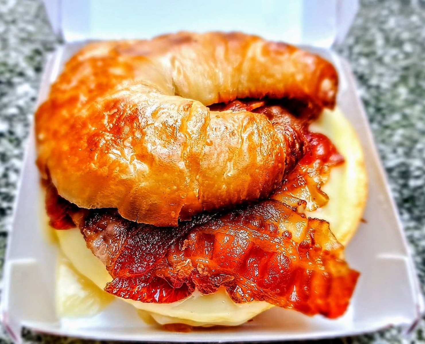 A close-up of a sandwich with crispy bacon and melted cheese inside a golden, flaky croissant, placed in a white takeaway box on a speckled gray surface.