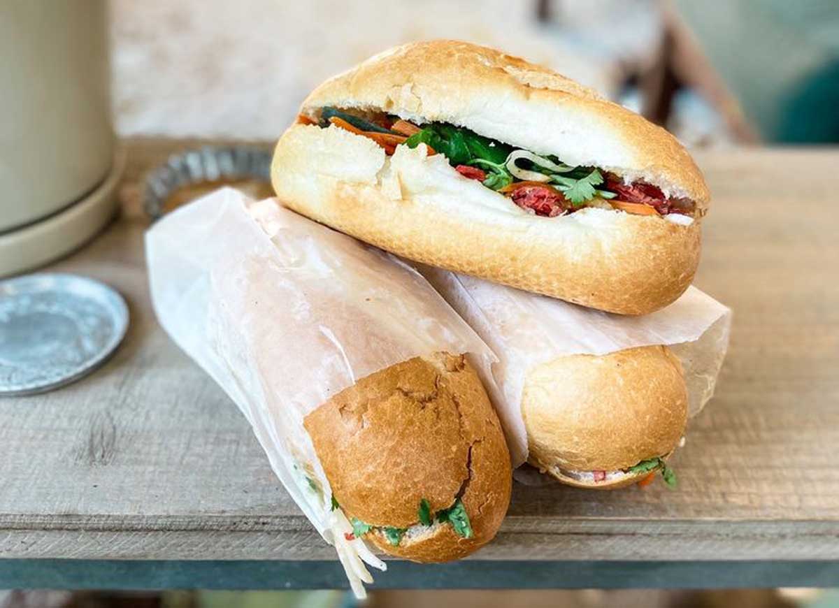 Three Vietnamese banh mi sandwiches on a wooden surface; two are wrapped in white paper, while one is unwrapped to reveal herbs, vegetables, and meats in a crispy baguette—a tasty find at cheap restaurants in every state.
