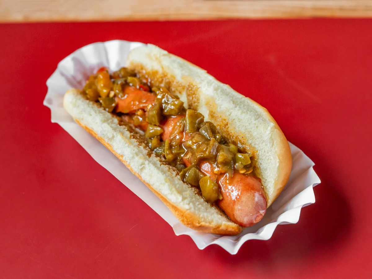 A hot dog in a white paper tray sits on a red surface. The sausage is topped with a chunky relish and sauce inside a soft bun.