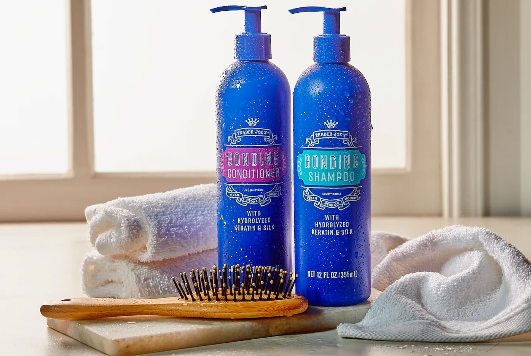 Two blue bottles of shampoo and conditioner labeled "Bonding Shampoo" and "Bonding Conditioner" sit on a counter next to a wooden hairbrush, a rolled white towel, and a folded white washcloth. A window is in the background.