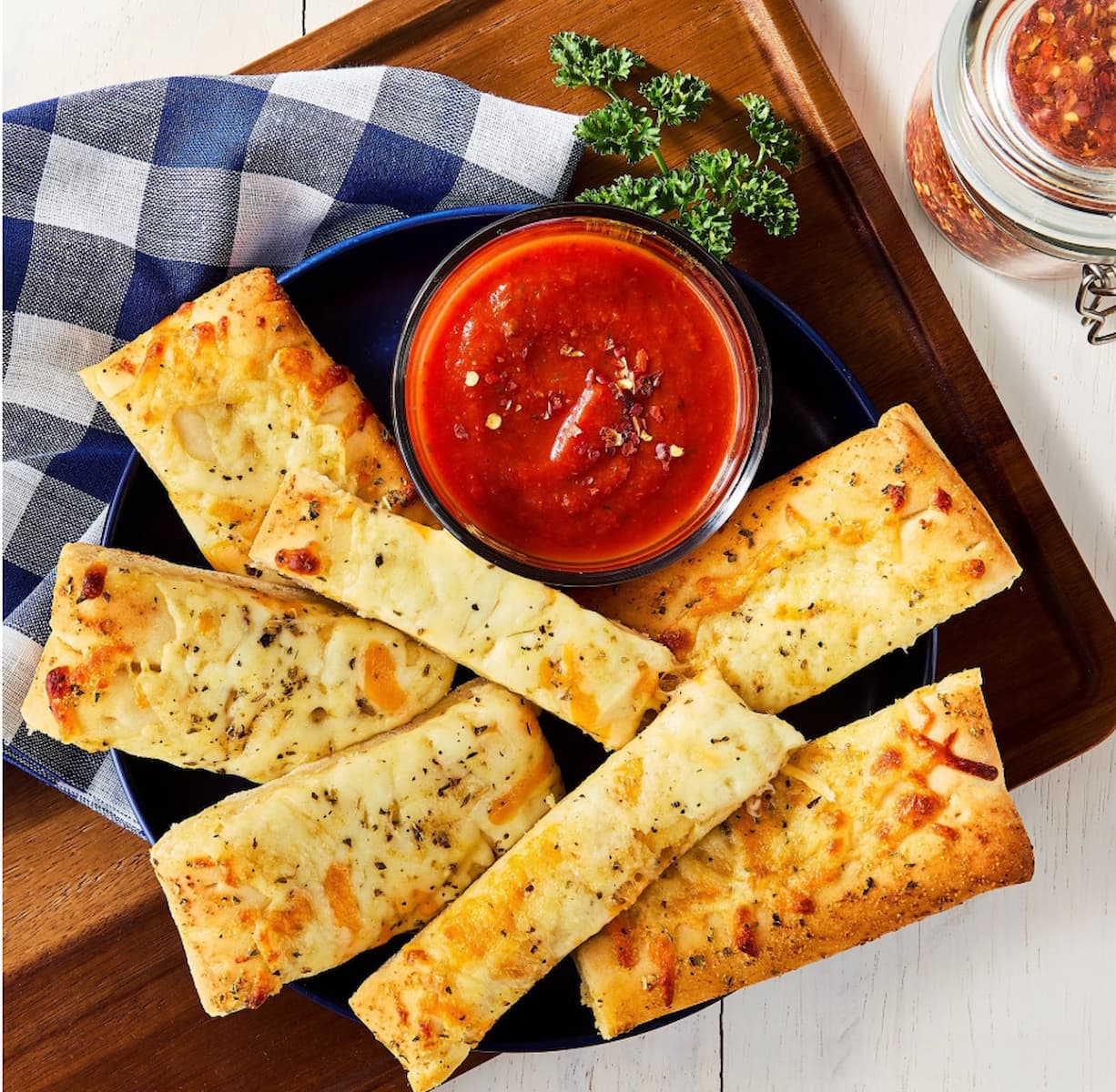 A plate of six cheesy breadsticks with herbs, served with a bowl of marinara sauce. A checkered napkin, parsley garnish, and a jar of red pepper flakes are nearby on a wooden surface.