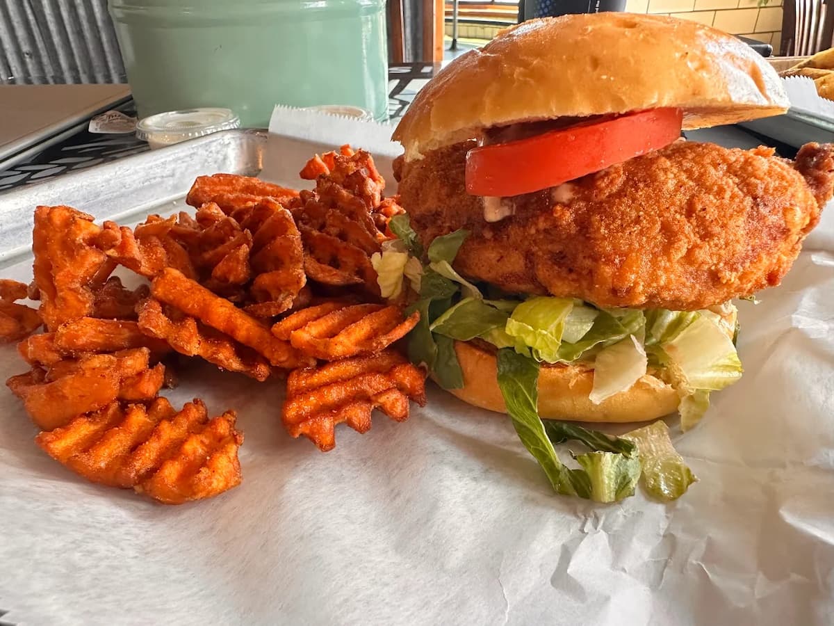 A breaded chicken sandwich with lettuce and tomato is served on a bun next to a pile of crispy waffle-cut sweet potato fries, offering a taste that could rival the best fried chicken in every state.