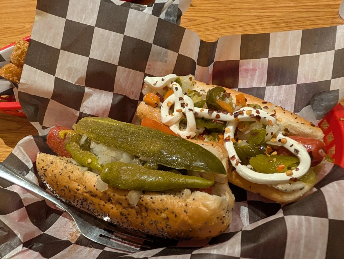 Two hot dogs in poppy seed buns, topped with relish, onions, pickles, peppers, jalapeños, mayonnaise, and chili flakes, are served on black-and-white checkered paper in a basket on a wooden table.