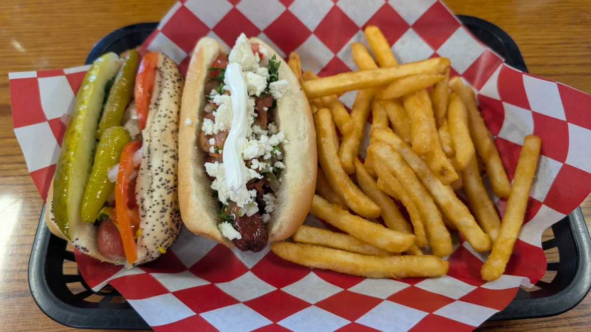 A food basket with a Chicago-style hot dog, a loaded hot dog topped with cheese and sour cream, and a serving of crinkle-cut french fries on red and white checkered paper.