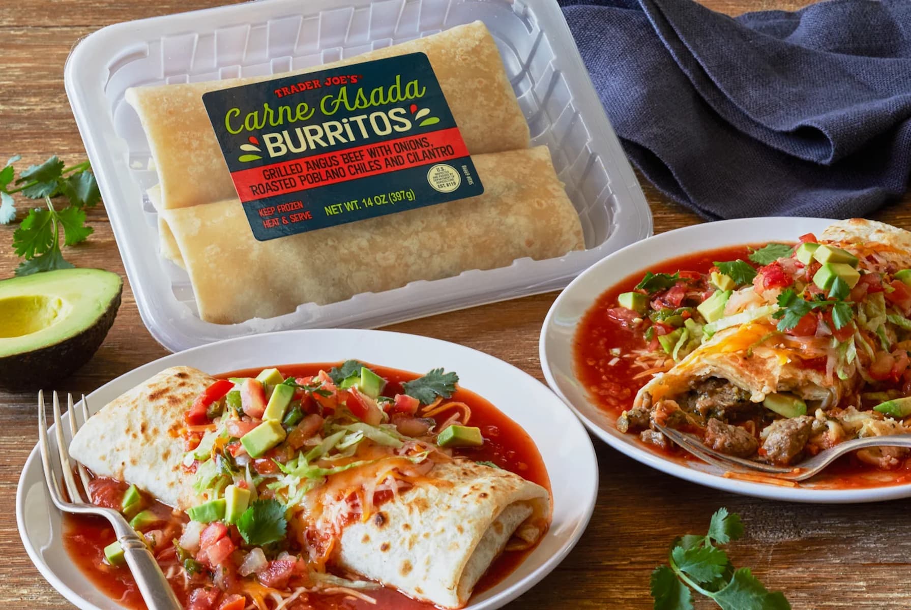 Two carne asada burritos on plates, topped with salsa, avocado, and cilantro, sit in front of a Trader Joe's package labeled "Carne Asada Burritos." An avocado half and herbs are nearby.
