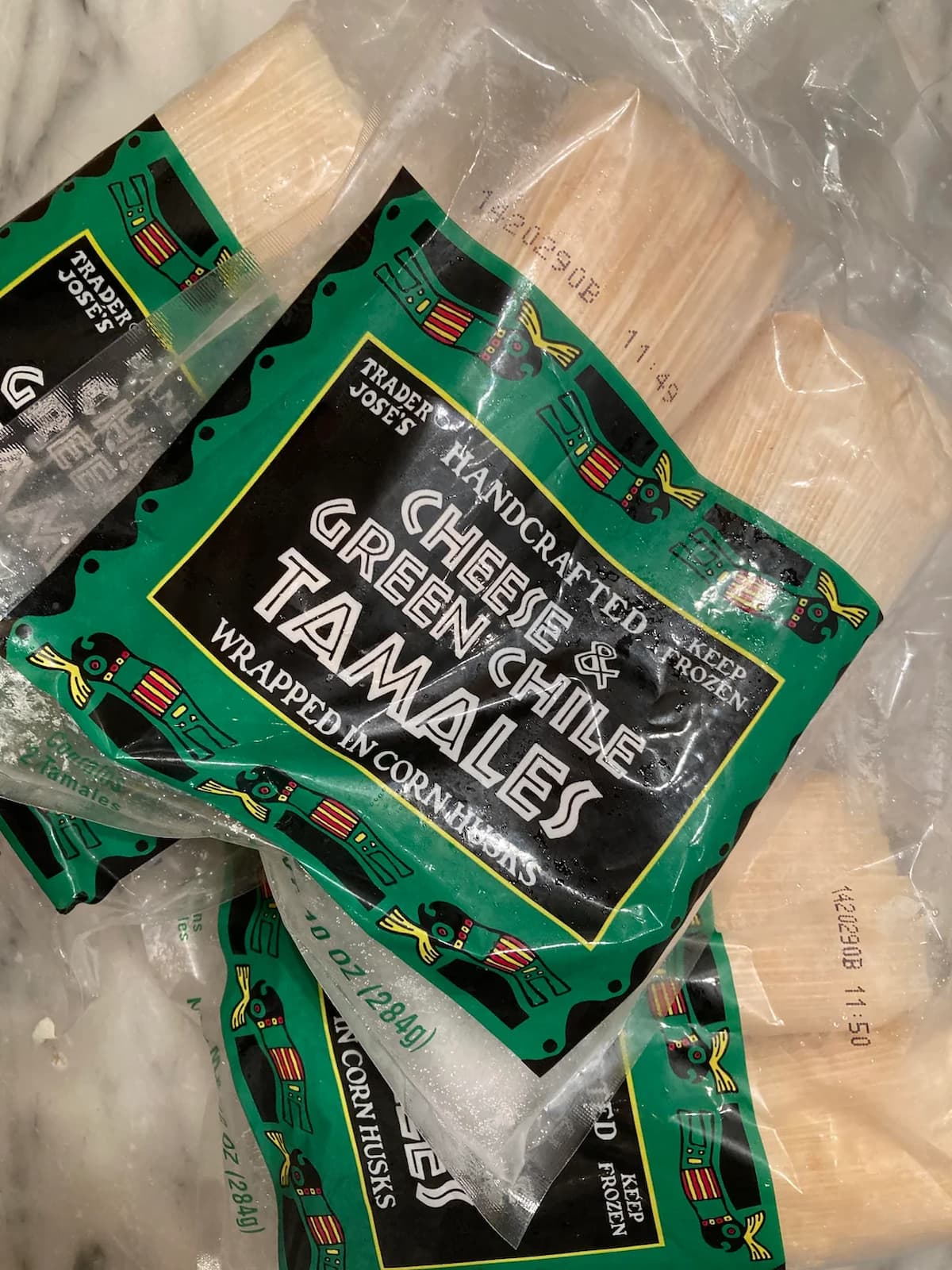 A package of Trader Joe’s Handcrafted Cheese & Green Chile Tamales wrapped in corn husks sits on a marble surface, with another partially visible package underneath.