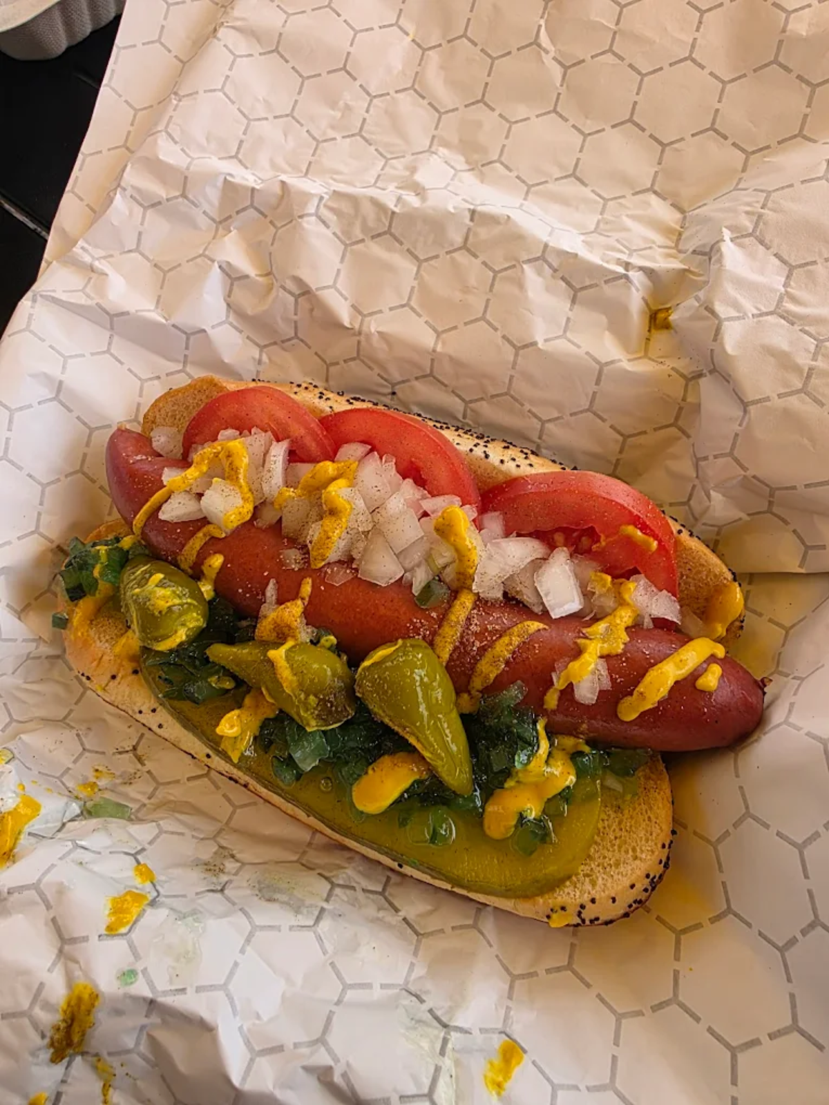 A Chicago-style hot dog on a poppy seed bun topped with tomato slices, chopped onions, relish, sport peppers, a pickle spear, mustard, and celery salt, served on white patterned paper.