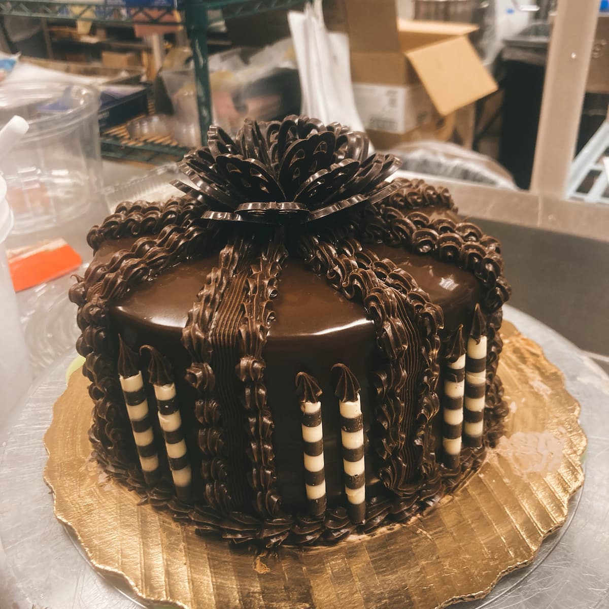 A round chocolate cake decorated with glossy chocolate ganache, piped chocolate designs, striped white and dark chocolate sticks, and a large chocolate flower on top, displayed on a gold tray.