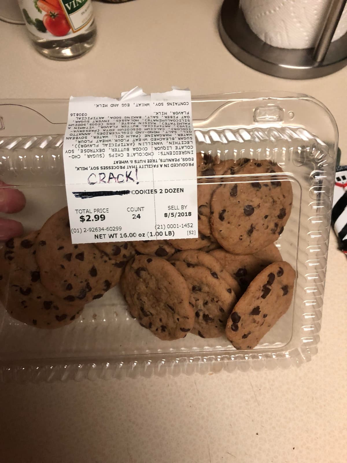 A clear plastic container holds chocolate chip cookies with a store label on top. The label shows the price, weight, and sell-by date. "CRACK!" is handwritten on the label in black marker.