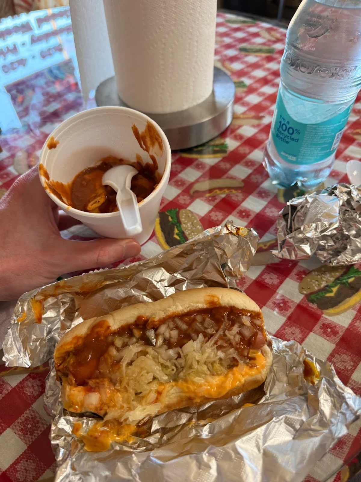 A hand holds a white cup of chili with a spoon, next to a chili cheese hot dog in foil on a red checkered tablecloth. There’s a bottle of water and a paper towel holder in the background.