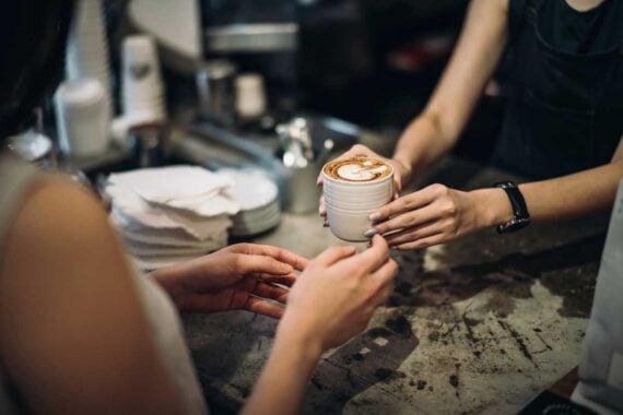 A barista hands a cup of coffee with latte art to a customer across a counter in a caf&eacute;; stacks of napkins and cups are visible in the background.