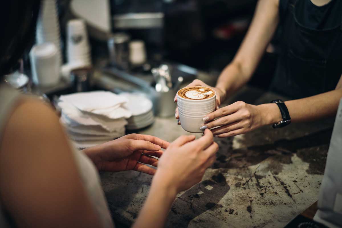 A barista hands a cup of coffee with latte art to a customer across a counter in a café; stacks of napkins and cups are visible in the background.
