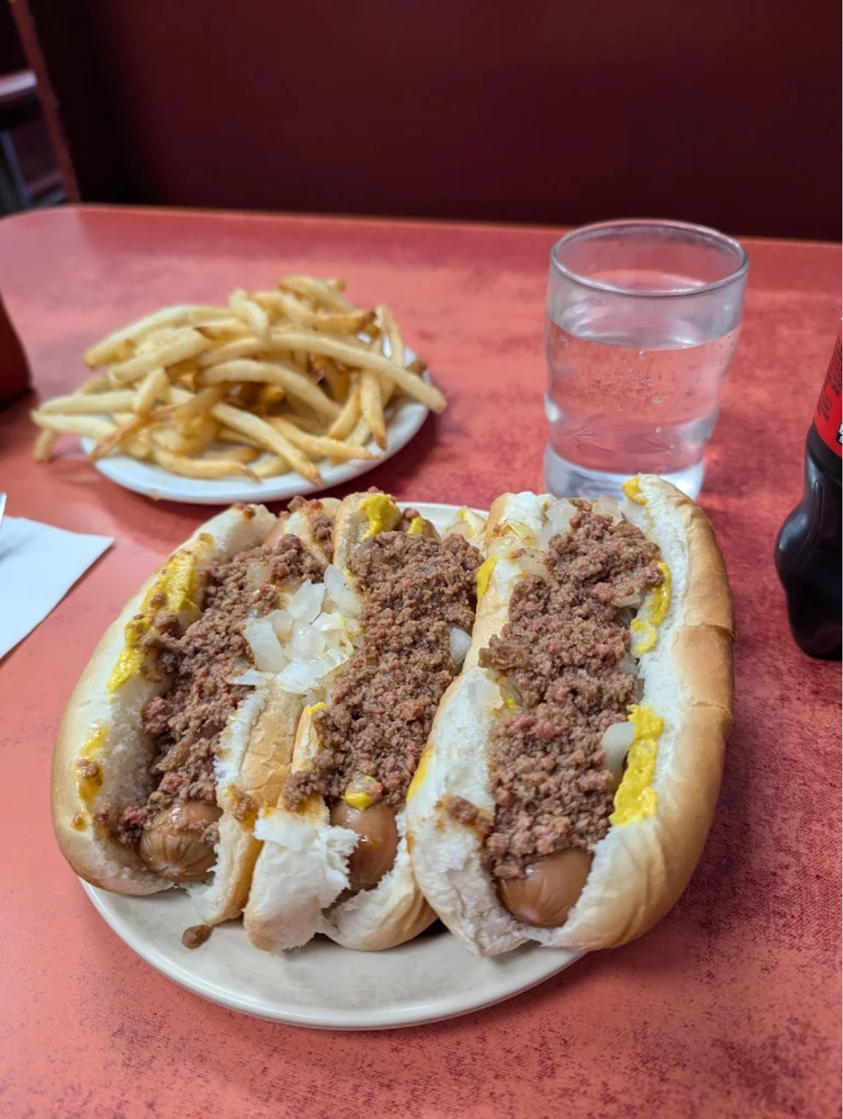 Three chili-topped hot dogs with mustard and chopped onions on a plate, served with a side of French fries, a glass of water, and a bottle of Coca-Cola on a red table.