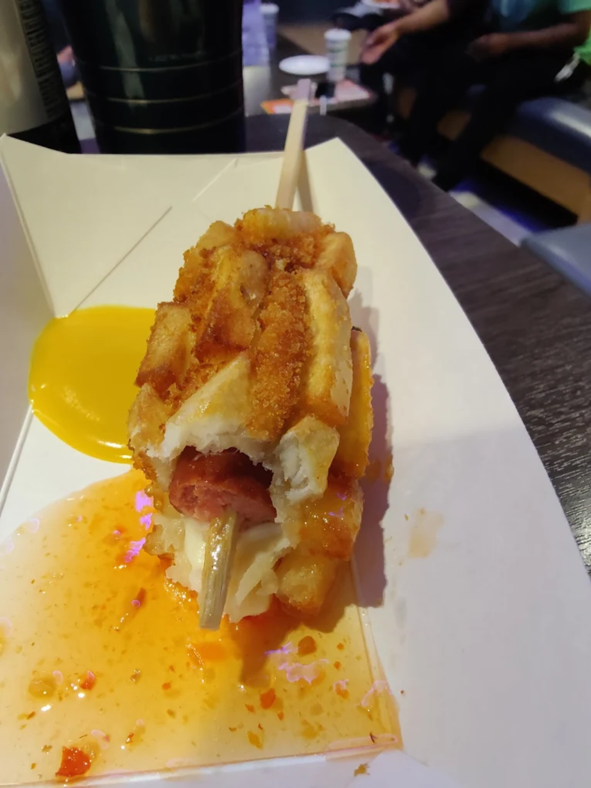 A close-up of a battered and fried corn dog on a stick, with waffle-like texture, partially eaten and served in a white tray with a yellow sauce and orange oil drips. Some people are blurred in the background.