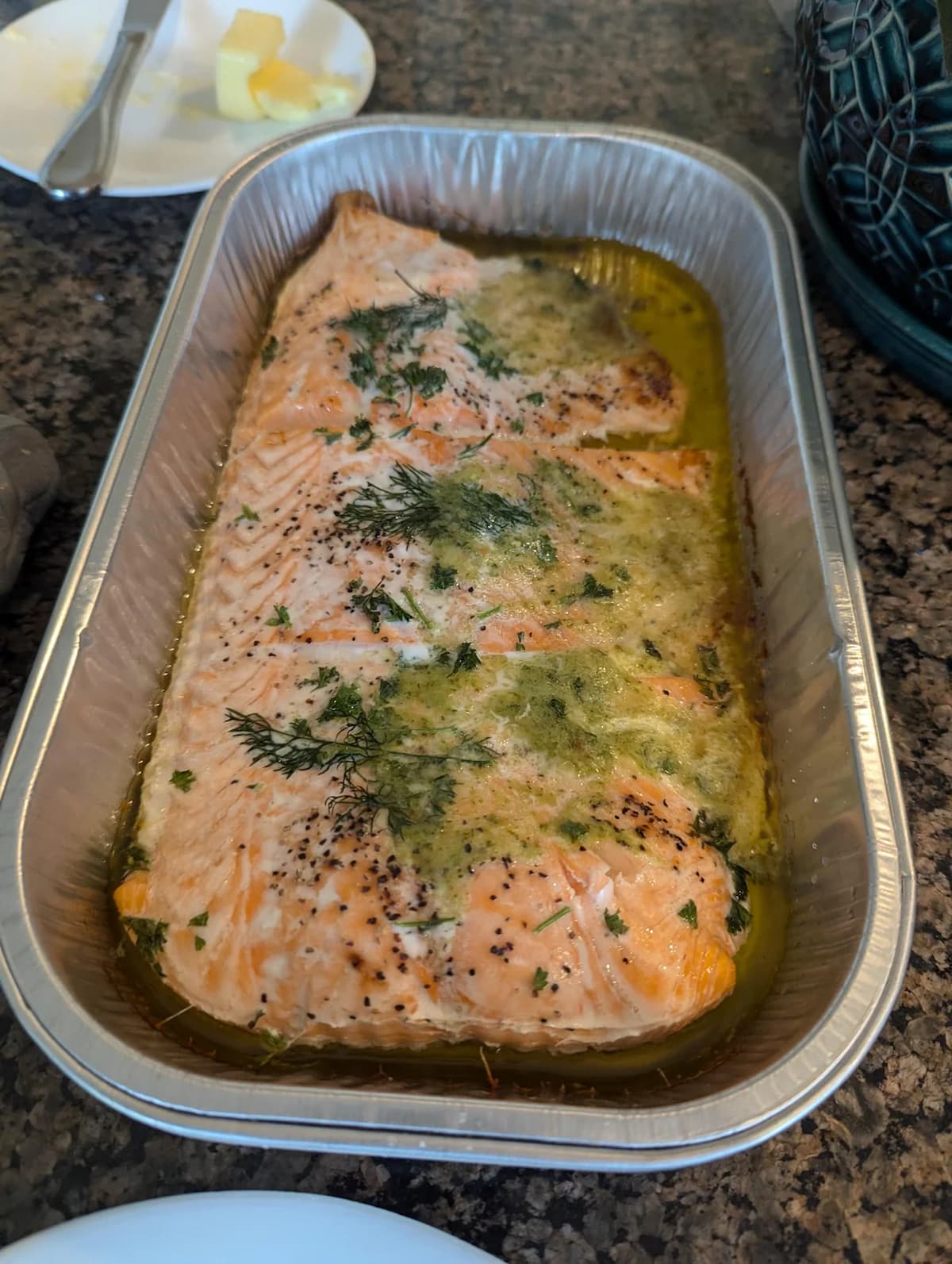 A baked salmon fillet garnished with herbs, sitting in a foil tray with melted butter and seasoning on a granite countertop. In the background, there’s a plate with butter and a knife.
