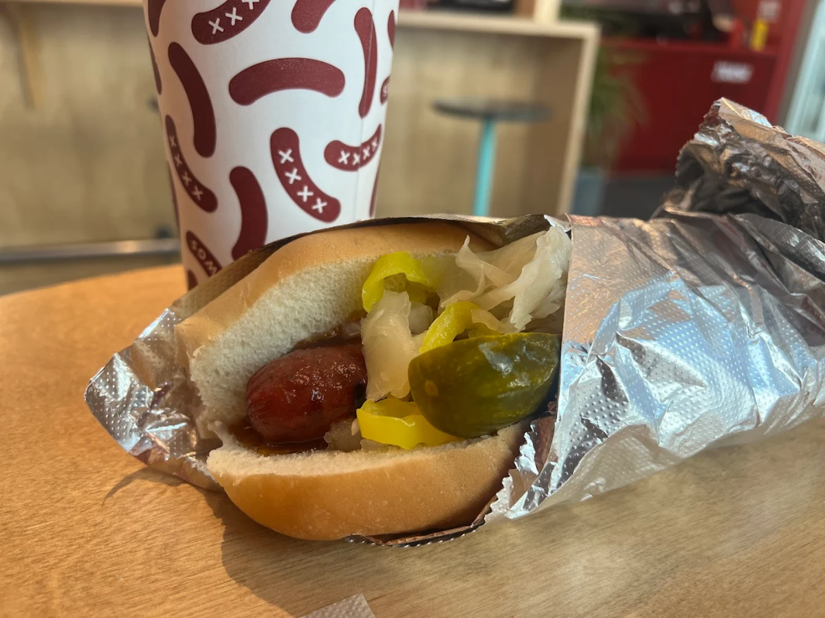 A hot dog in a bun with pickles, sauerkraut, and sliced peppers, partially wrapped in foil, sits on a table next to a paper cup with a red and white sausage pattern.