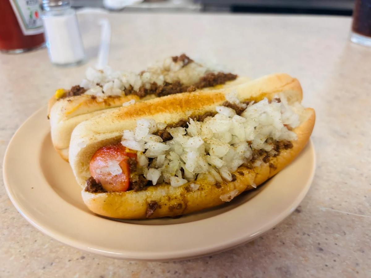 Two chili dogs topped with chopped onions sit on a beige plate on a countertop. The background includes a salt shaker, a straw, and a glass of dark soda.