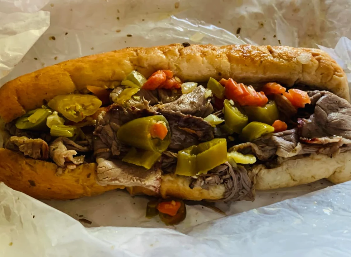 A close-up of an Italian beef sandwich on a white wrapper, filled with sliced roast beef and topped with chopped green peppers and red peppers.