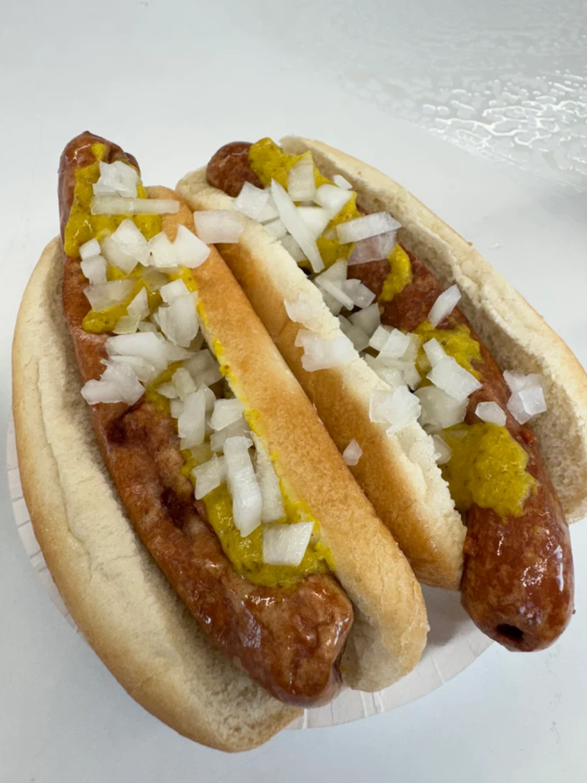 Two grilled hot dogs in buns topped with chopped onions and yellow mustard, served on a white plate with a light background.
