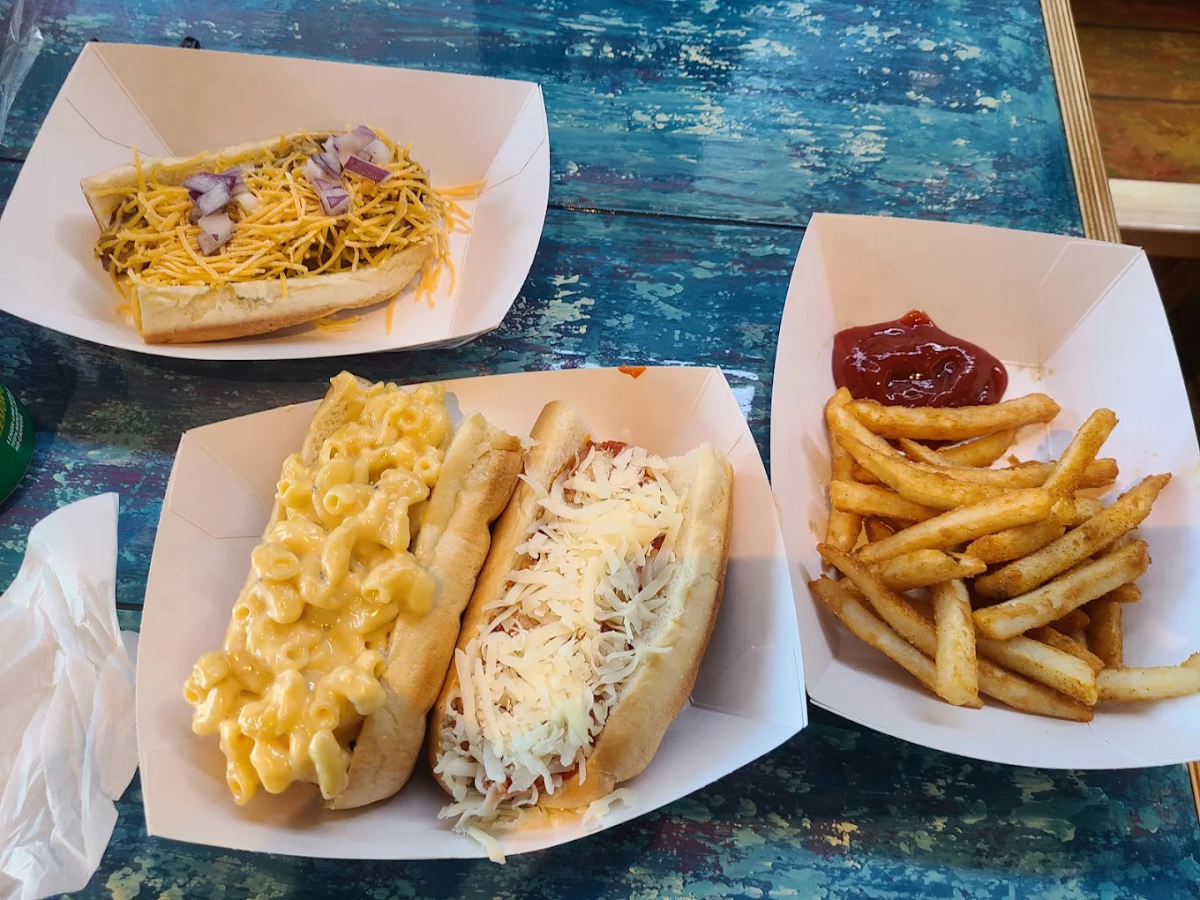 Three paper trays on a blue rustic table: one holds a hot dog with shredded cheese and onions, another holds fries with ketchup, and the third has two hot dogs—one topped with macaroni and cheese, the other with shredded cheese.