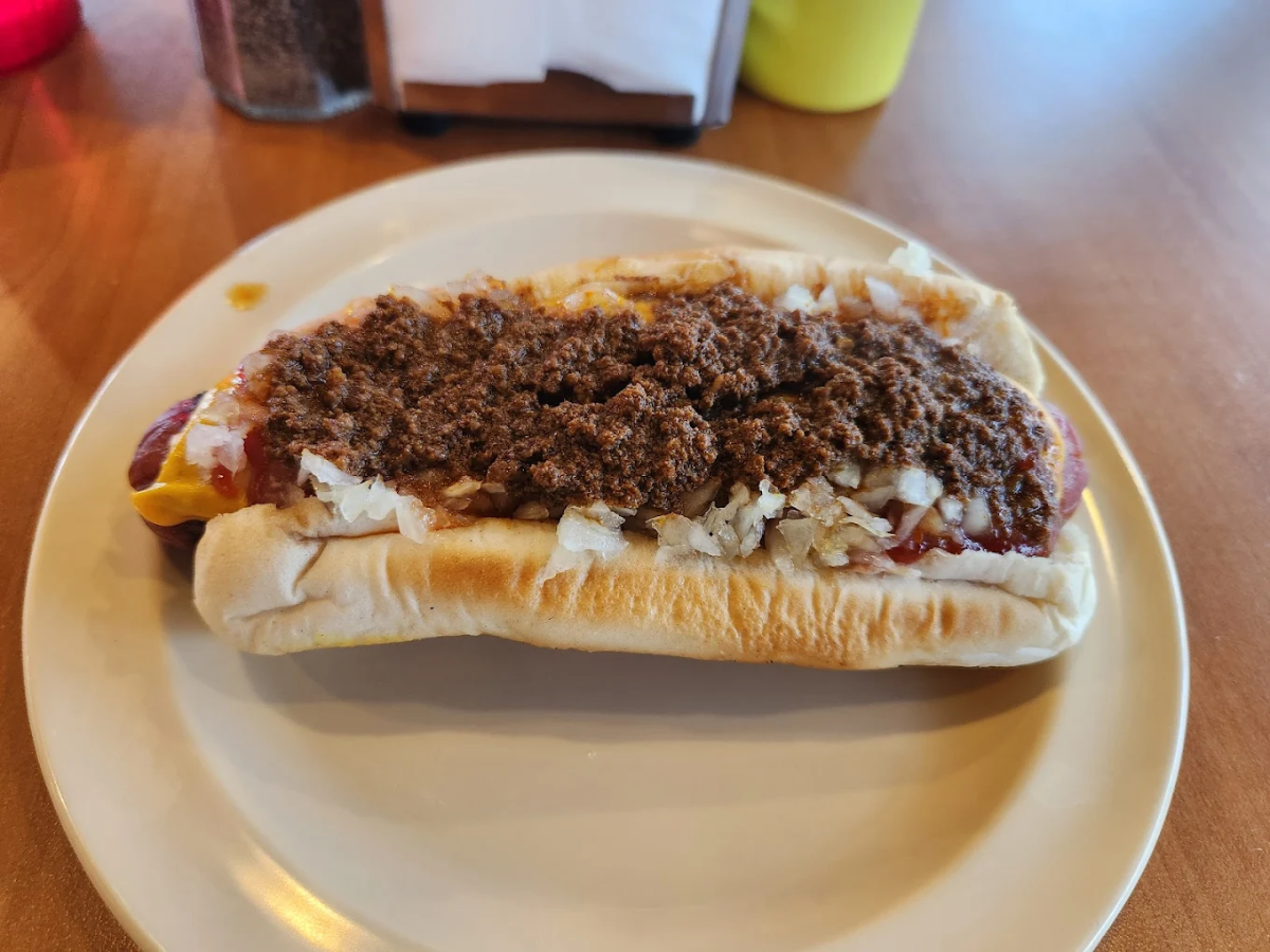 A hot dog on a white plate topped with chopped onions, mustard, and a generous layer of chili, sitting on a wooden table with condiments in the background.