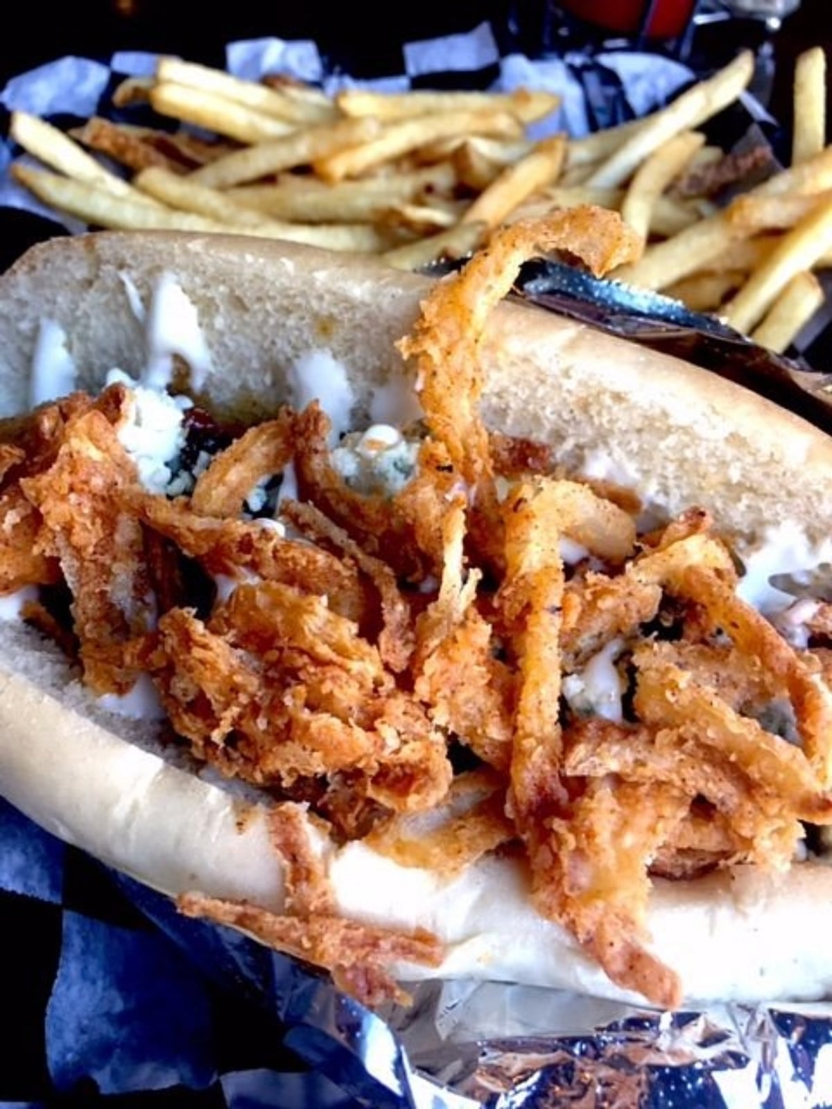 A close-up of a sandwich filled with crispy fried onions and drizzled with white sauce, served on a hoagie roll with French fries in the background.