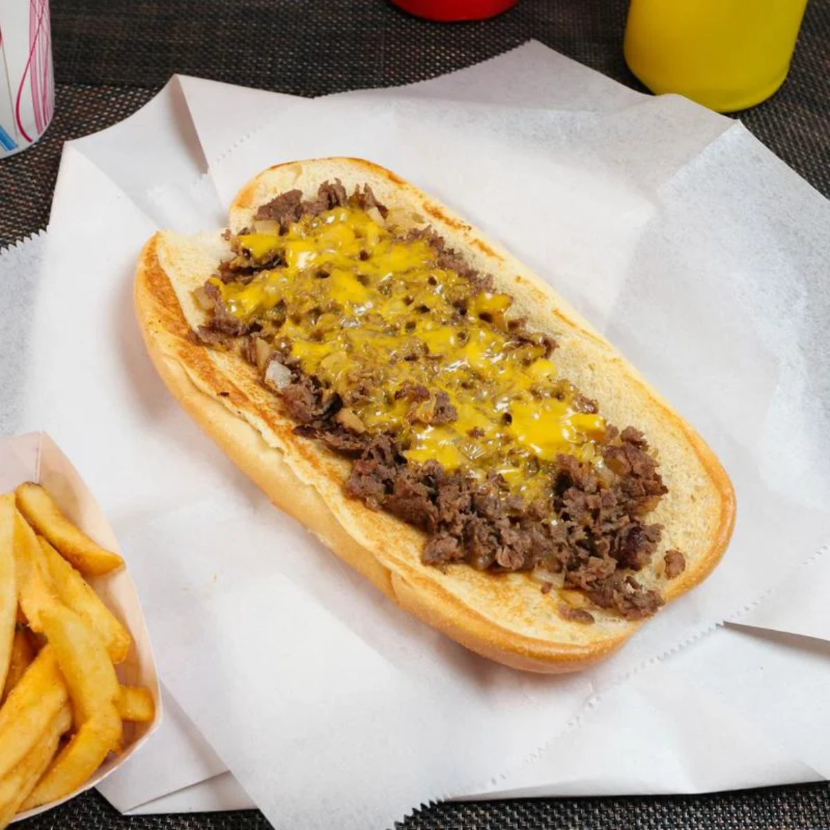 A hoagie roll filled with chopped steak and melted cheddar cheese sits on parchment paper next to a container of crinkle-cut French fries. Bottles of ketchup and mustard are in the background.