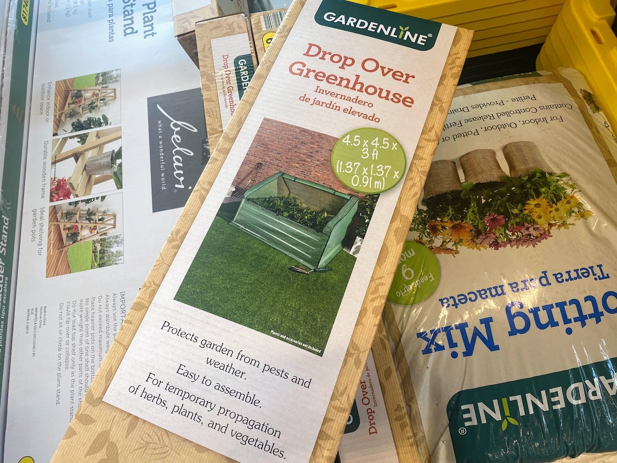 A Drop Over Greenhouse kit by Gardenline is displayed in a store, showing images of the assembled greenhouse on grass. The packaging highlights features like pest protection and easy assembly for temporary plant use.