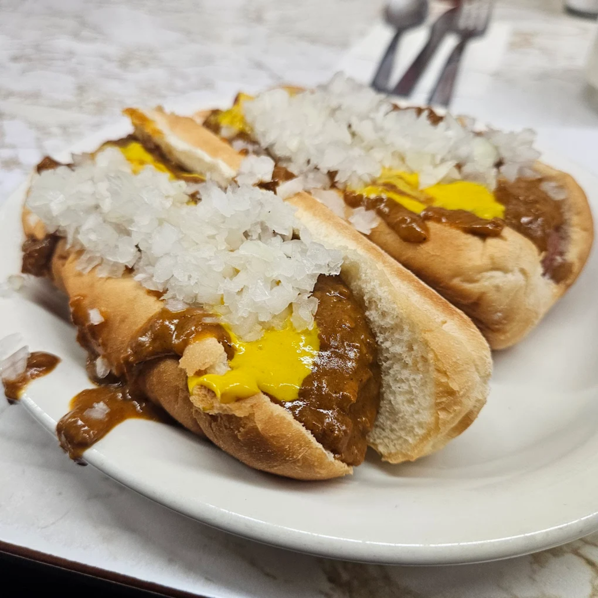 Two hot dogs on a white plate topped with chili, yellow mustard, and a generous amount of chopped onions, with metal utensils in the background on a marble table.