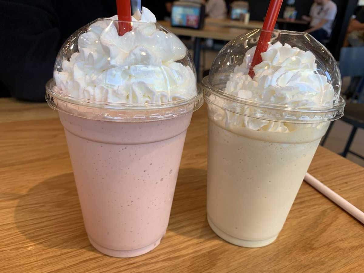 Two milkshakes with whipped cream and red straws sit on a wooden table. One milkshake is pink, likely strawberry, and the other is light yellow, possibly vanilla or banana. Both are in clear plastic cups with dome lids.