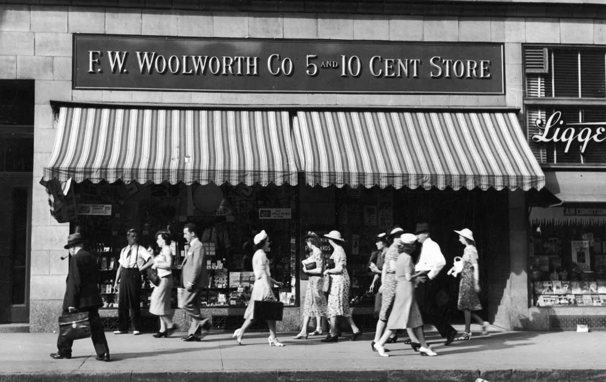 Black and white photo of people walking past an F.W. Woolworth Co. 5 and 10 Cent Store; storefront has striped awning and displays goods in windows; men and women wear 1940s-style clothing.