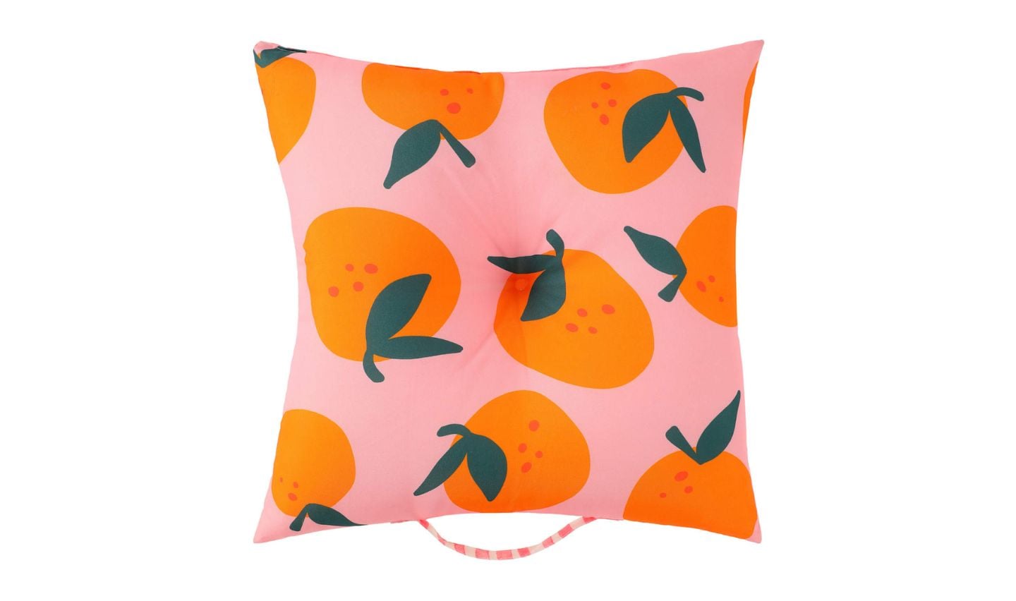 A square pillow with a pink background featuring a pattern of large orange fruit, green leaves, and small red dots. Part of the new Five Below products, it has a button tuft in the center and a pink-and-white striped handle at the bottom.