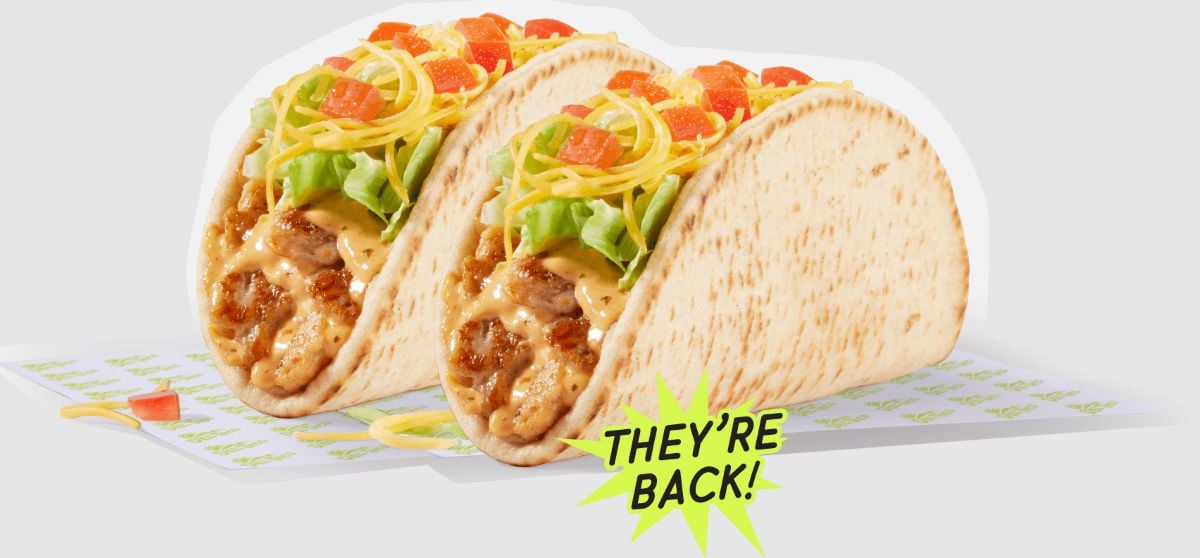 Two flatbread tacos from the new fast-food menu, filled with seasoned beef, shredded cheese, lettuce, diced tomatoes, and a creamy sauce. A green burst graphic announces "THEY'RE BACK!" in front of the tacos.