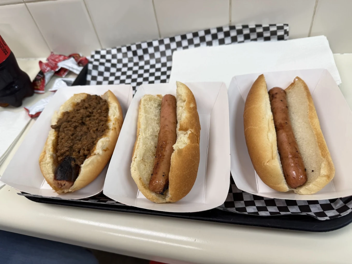 Three hot dogs in buns are placed on a tray with a checkered paper liner; one has chili on top, while the others are plain. There are napkins and condiment packets in the background.