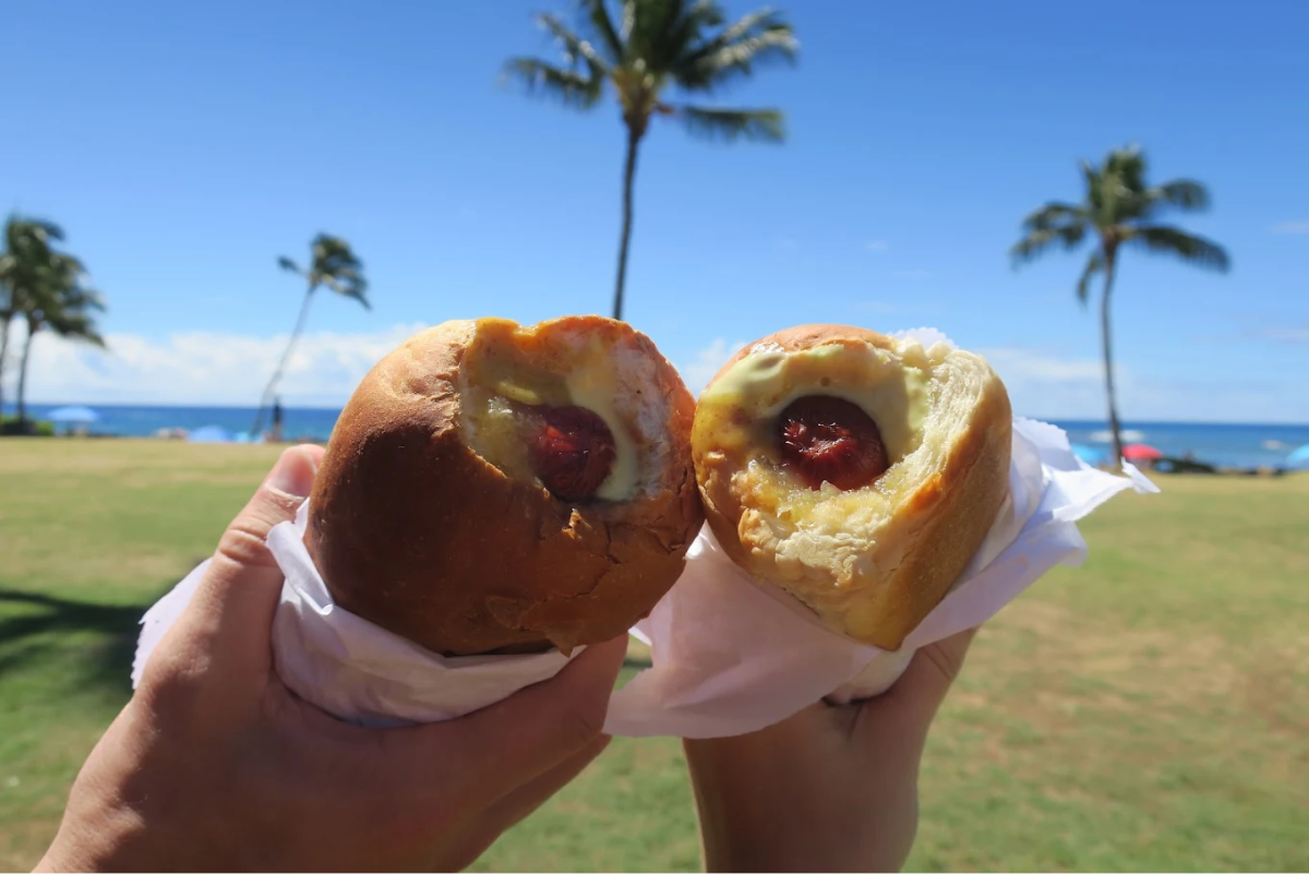 Two hands hold up Hawaiian-style hot dog buns with sausage and cheese inside, against a backdrop of palm trees, a grassy park, and the ocean under a bright blue sky.