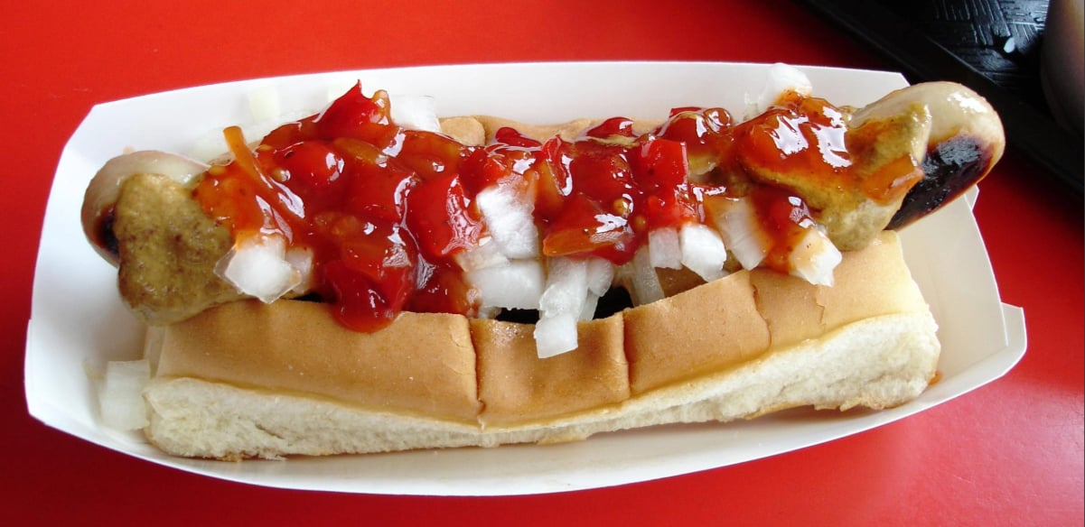 A cooked sausage in a bun topped with chopped onions, ketchup, and mustard, served in a white paper tray on a red surface.