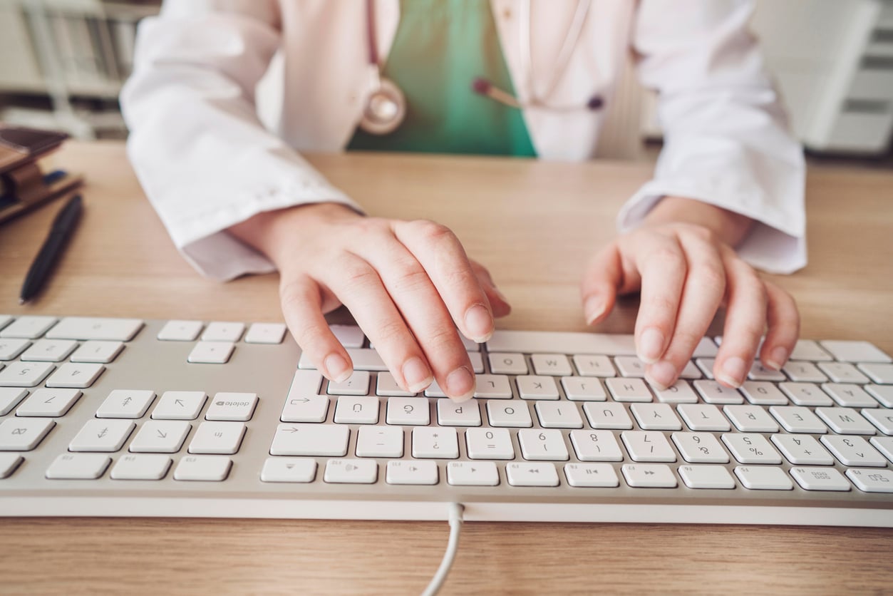A person wearing a white coat types on a computer keyboard at a desk, with a stethoscope visible around their neck, suggesting they are a healthcare professional.