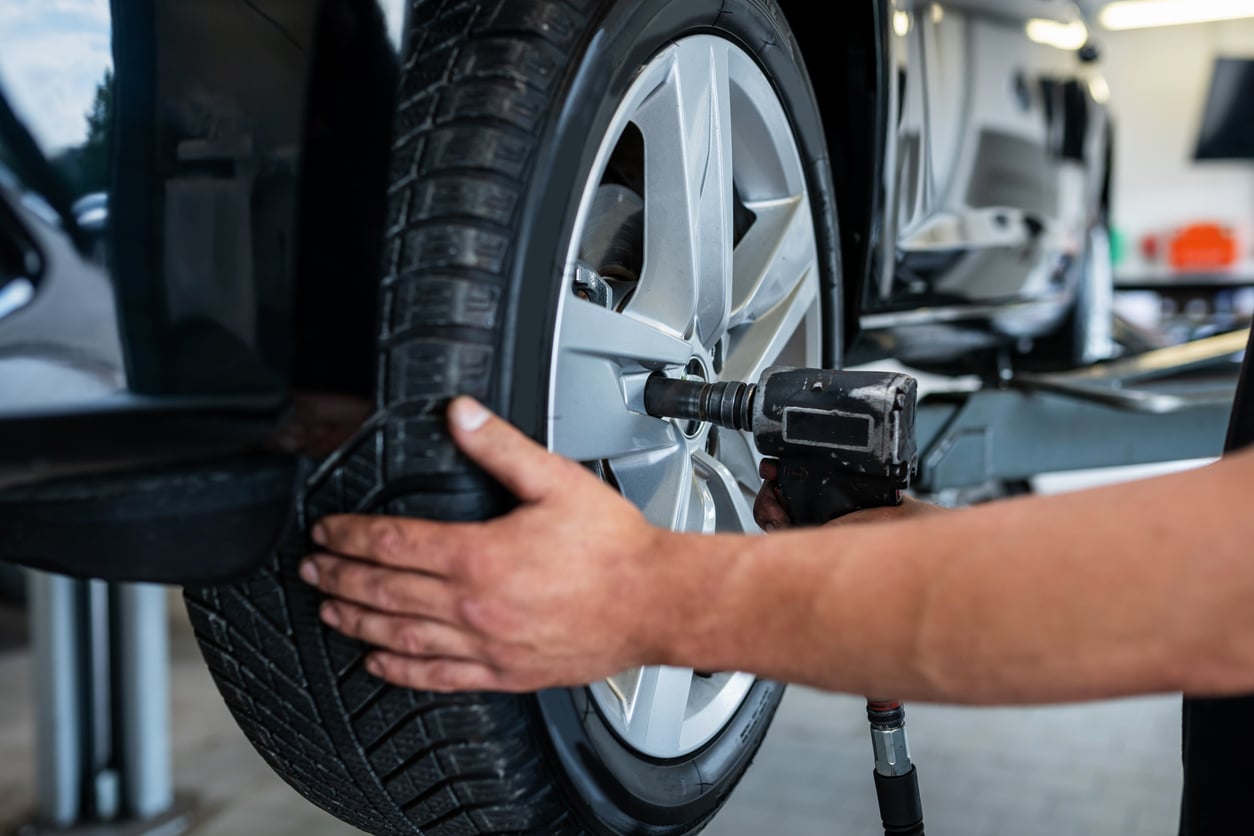 A mechanic uses a power tool to tighten the lug nuts on a car wheel, with both hands steadying the tire. The car is lifted on a service platform in an auto repair shop.