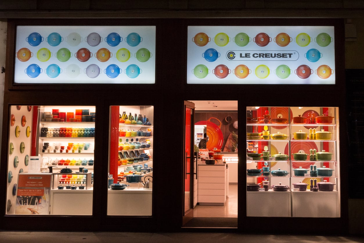 Storefront of a Le Creuset shop at night, featuring brightly lit displays of colorful cookware, dishes, and kitchen accessories arranged on shelves in the windows. The store sign and decorative plates are visible above the entrance.