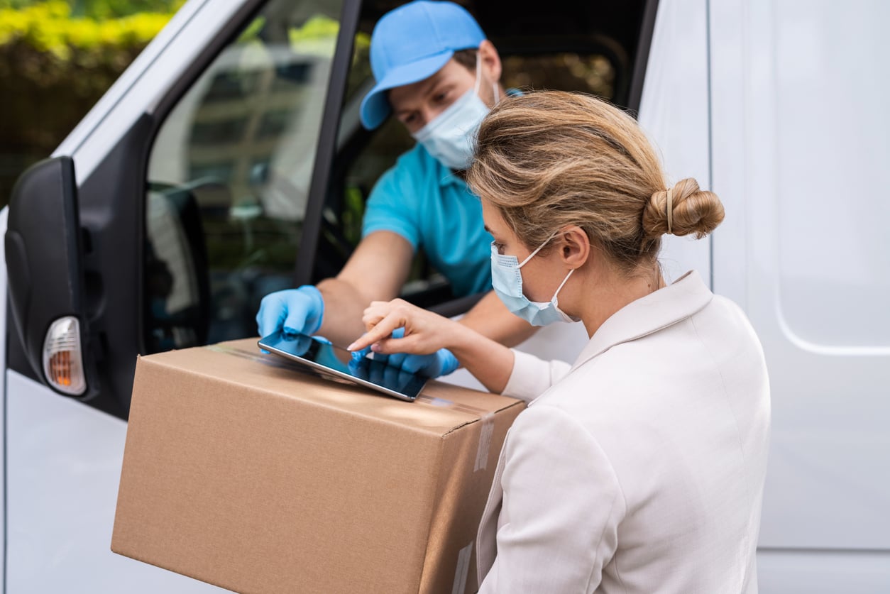 A woman wearing a mask receives a package from a courier in a blue cap and mask, signing for the delivery on a digital tablet next to a white van.