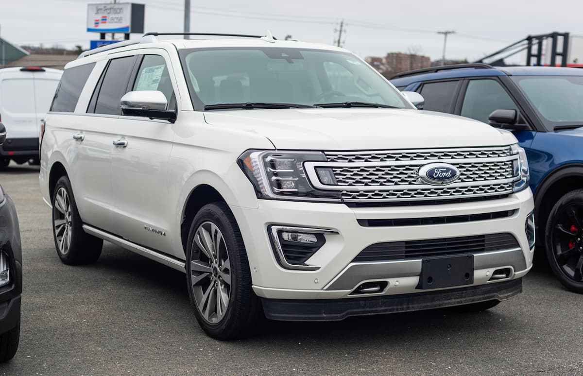 A white Ford Expedition SUV is parked at a car dealership, facing slightly left. The vehicle has chrome accents, a large front grille with the Ford logo, and alloy wheels—shown amid recent ford recall news. Other cars are partially visible nearby.