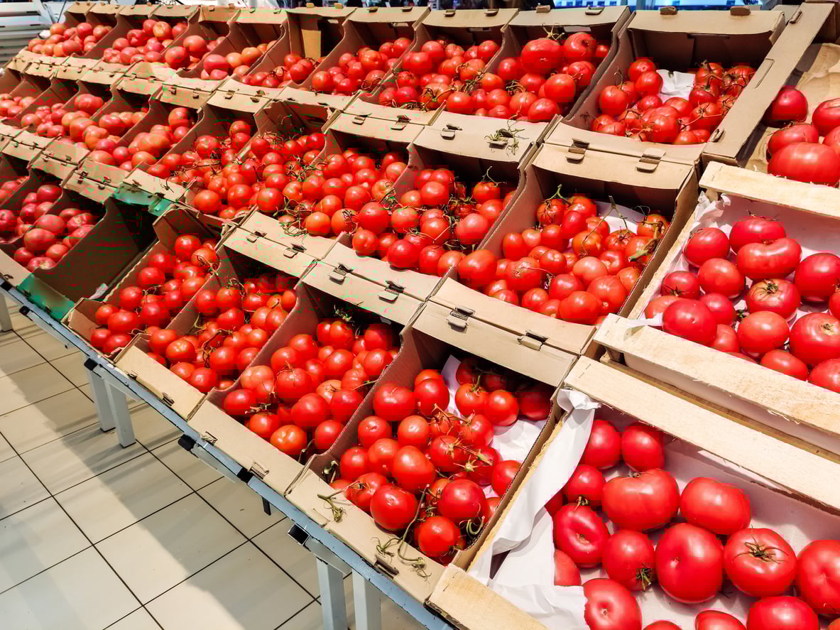 Display of fresh red tomatoes in cardboard boxes on angled supermarket shelves, arranged in neat rows. The tomatoes vary in size and shape, and the tiled floor is visible in the foreground.