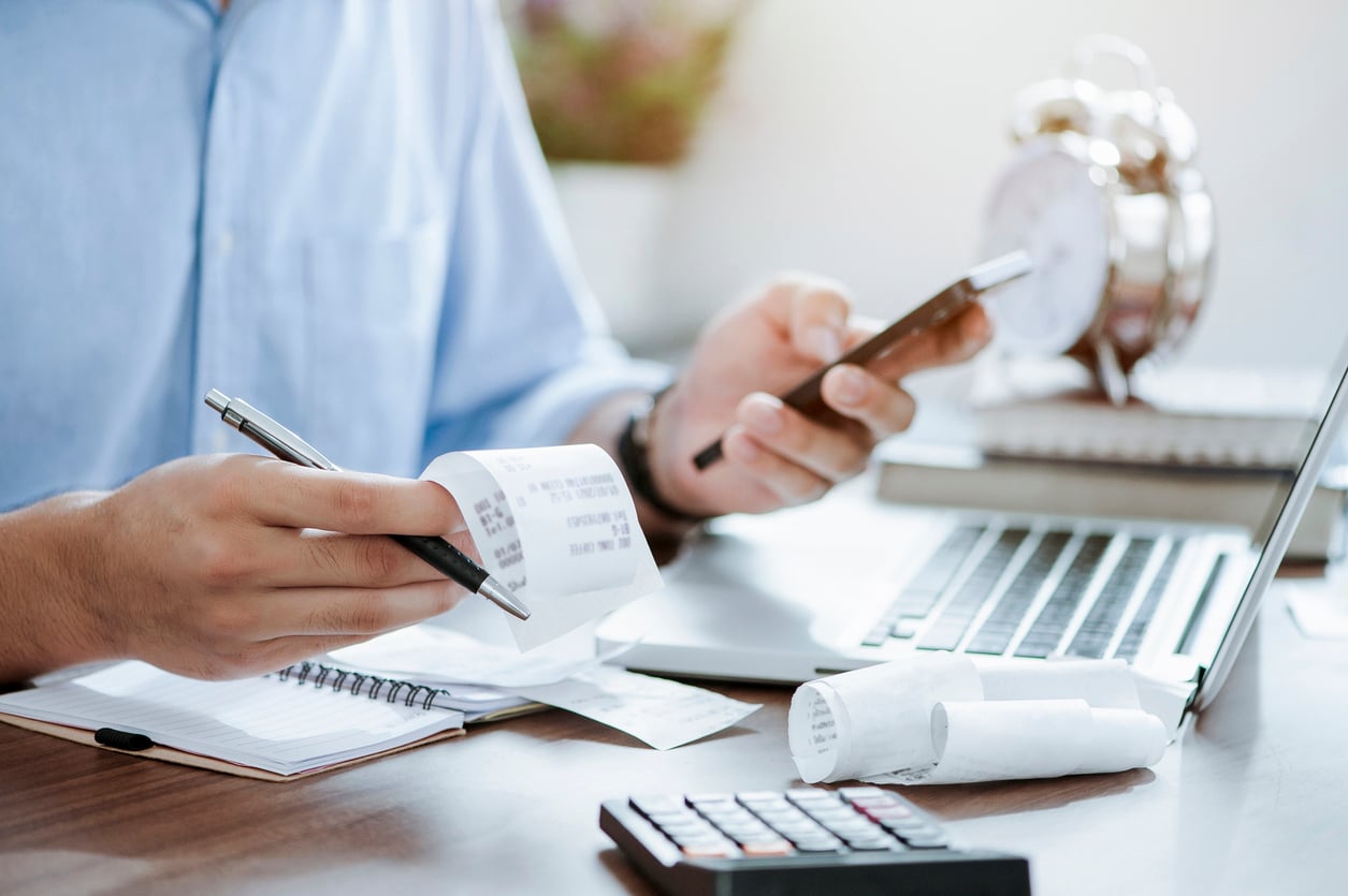 A person sits at a desk with a laptop, calculator, pen, and receipts, holding a smartphone in one hand and examining a receipt with the other, suggesting they are managing finances or budgeting.