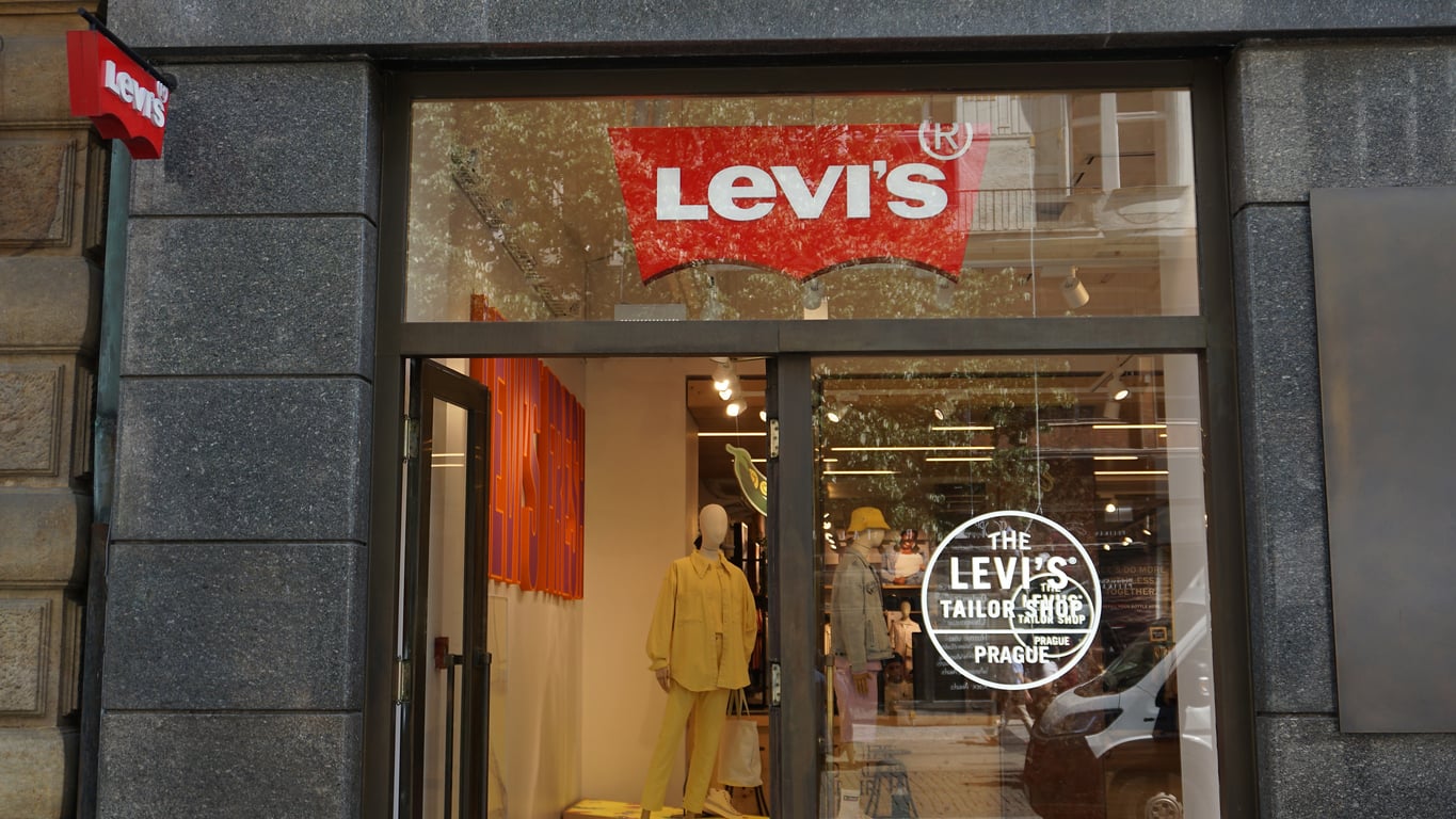 A Levi’s store entrance with a large red Levi’s logo above the glass door. Inside, a mannequin in yellow clothing is visible, and the store's name and logo are printed on the glass. Trees and buildings reflect in the window.