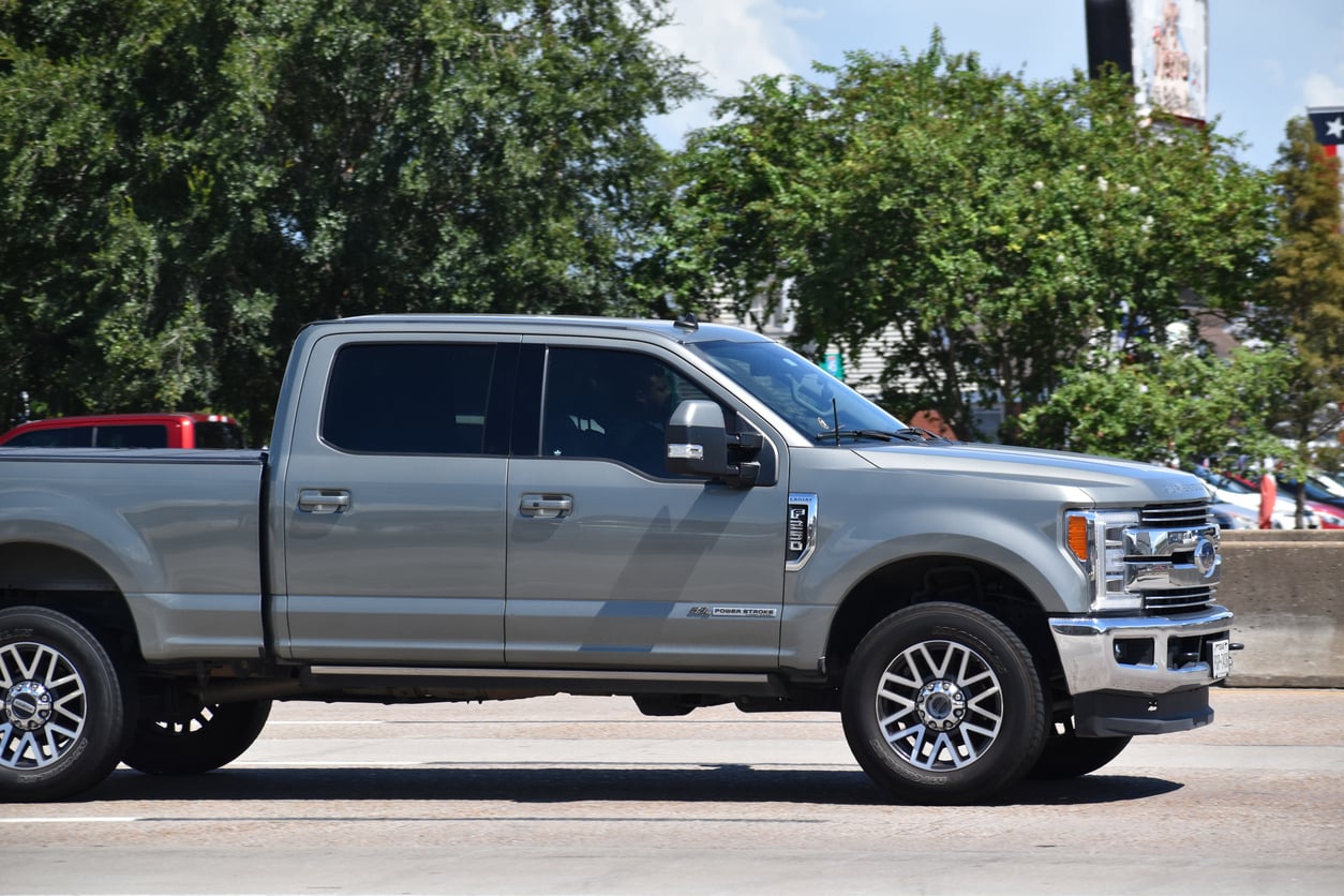 A silver Ford pickup truck with tinted windows drives on a road beside a concrete barrier, with trees and a partly cloudy sky in the background.