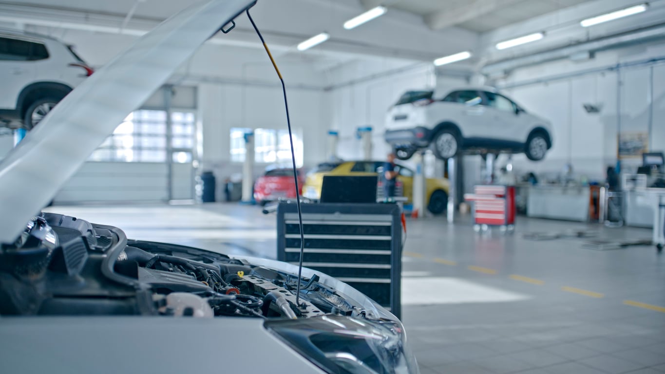 A car repair garage with several vehicles inside; one car in the foreground has its hood open, and another car is raised on a hydraulic lift. Tools and equipment are visible around the well-lit, spacious workshop.