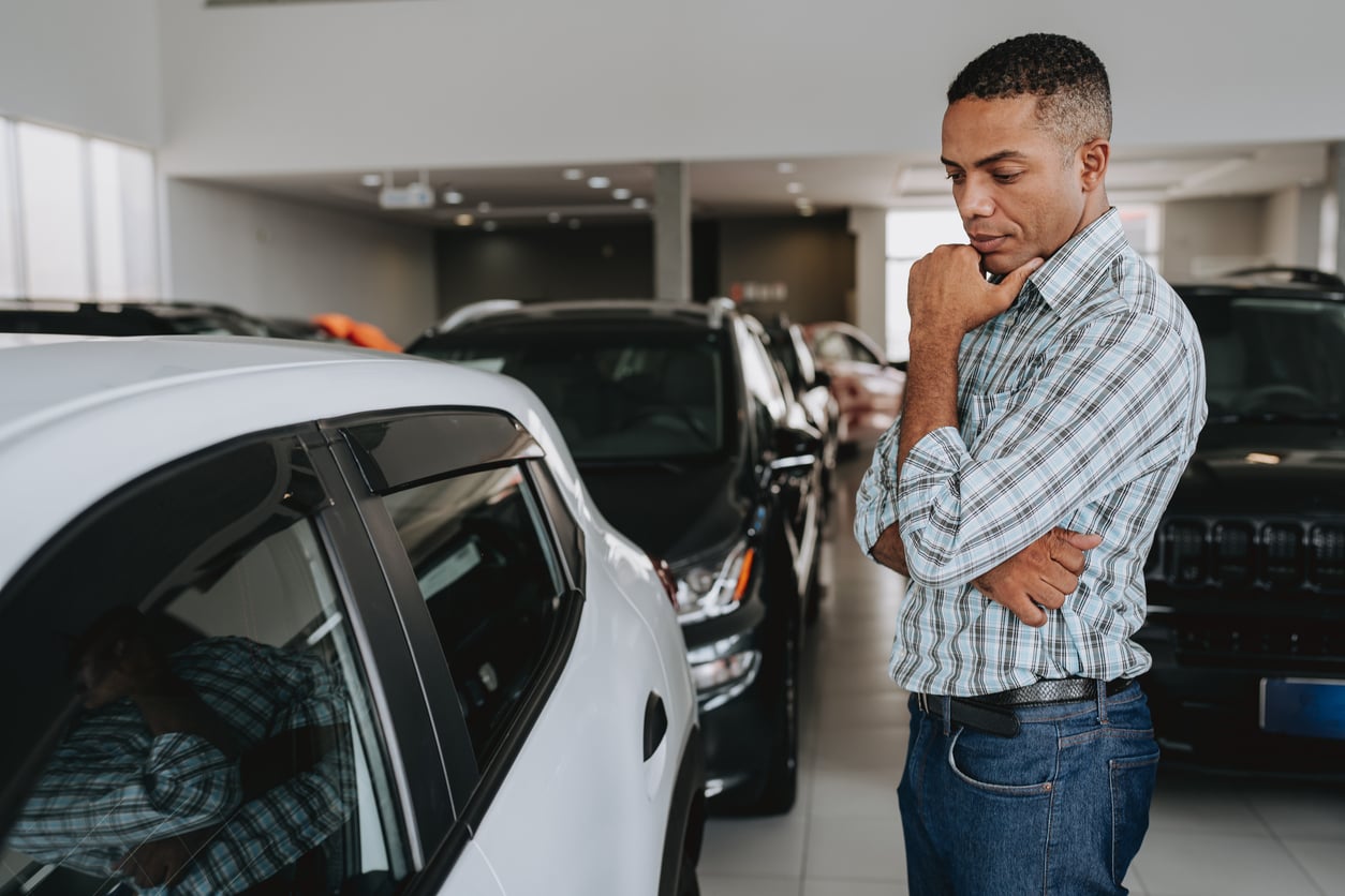 A man in a plaid shirt stands in a car showroom, thoughtfully looking at a white car among several parked vehicles, with his hand resting on his chin.