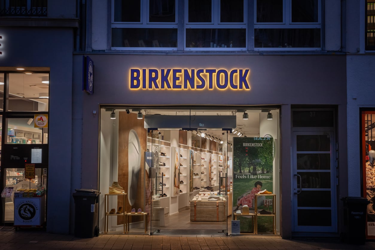 Storefront of a Birkenstock shoe store at night, brightly lit inside with shelves displaying sandals and shoes, large brand sign above entrance, and a poster featuring footwear in the window.