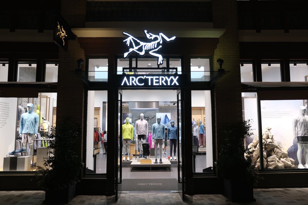 Storefront of Arc'teryx at night, with a brightly lit logo above the entrance. Mannequins in athletic outdoor clothing are visible through large glass windows and door.