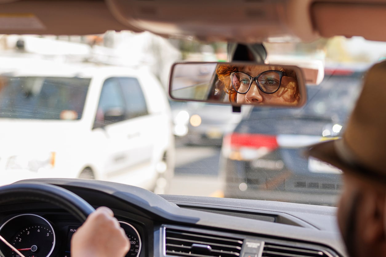 A person driving a car is seen from behind, with their hand on the steering wheel. Their face, wearing glasses, is reflected in the rearview mirror, and cars are visible through the windshield in traffic.